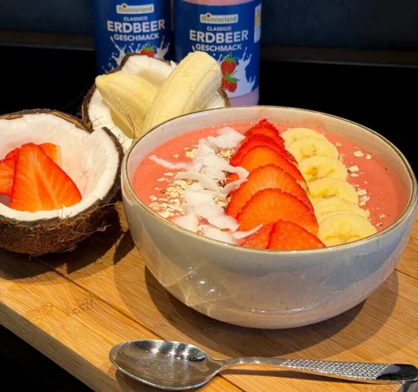 Smoothie Bowl mit Erdbeeren, Bananenscheiben und Kokosraspeln, dahinter eine halbe Kokosnuss